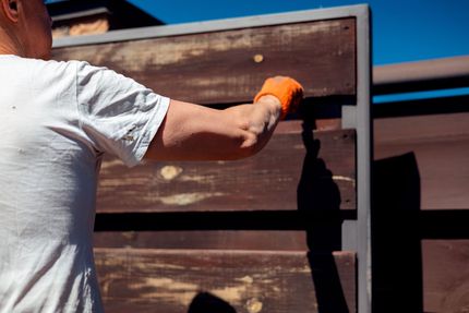 Person in orange gloves painting a wooden fence brown outdoors in bright sunlight