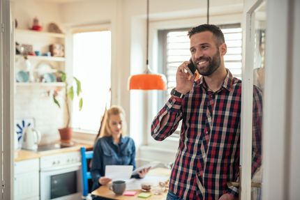 Man speaking on a phone in a bright kitchen while a woman works at a table in the background