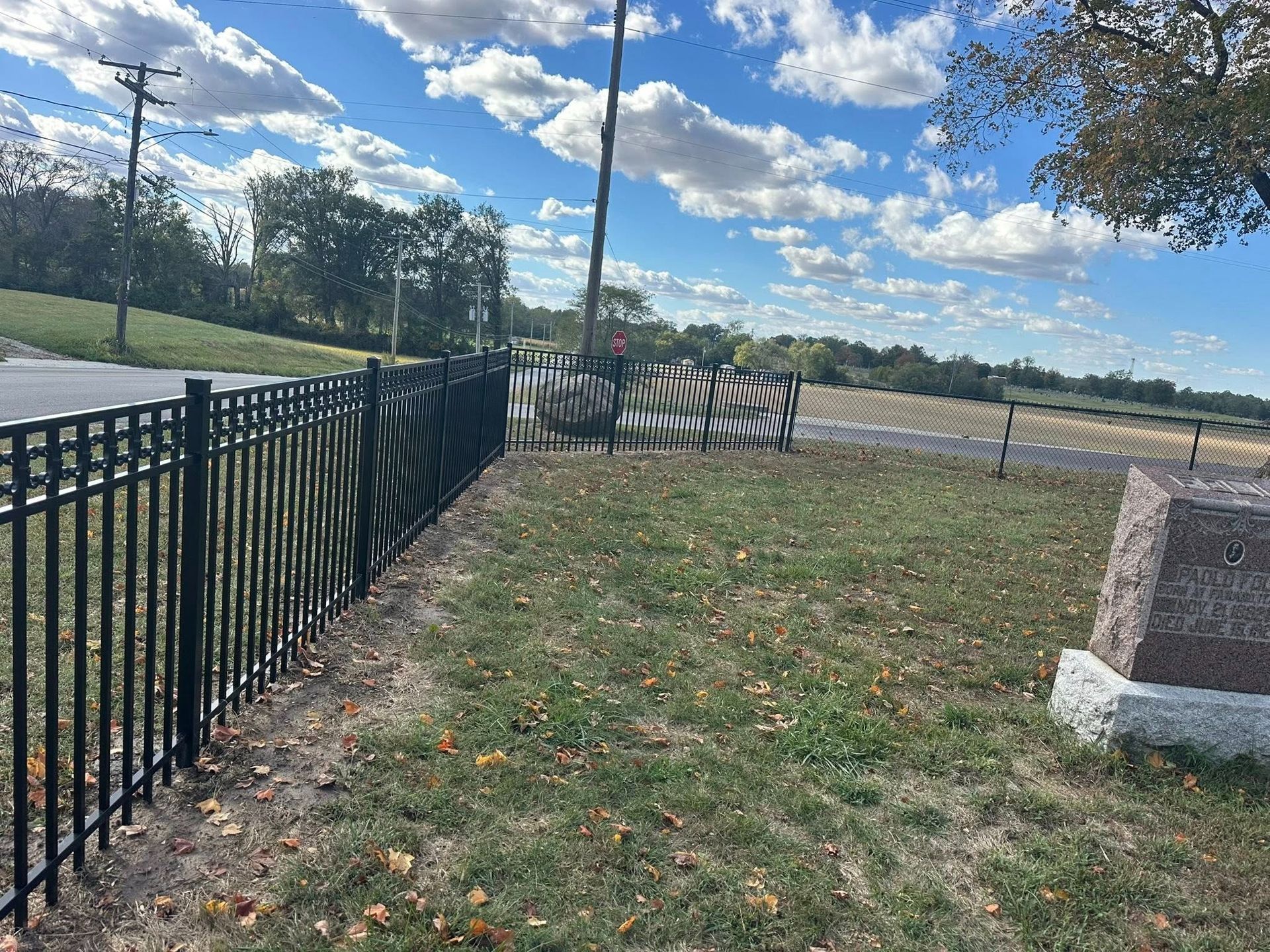 Grassy lot beside a black metal fence under a blue sky with clouds, near a road and stone pillar
