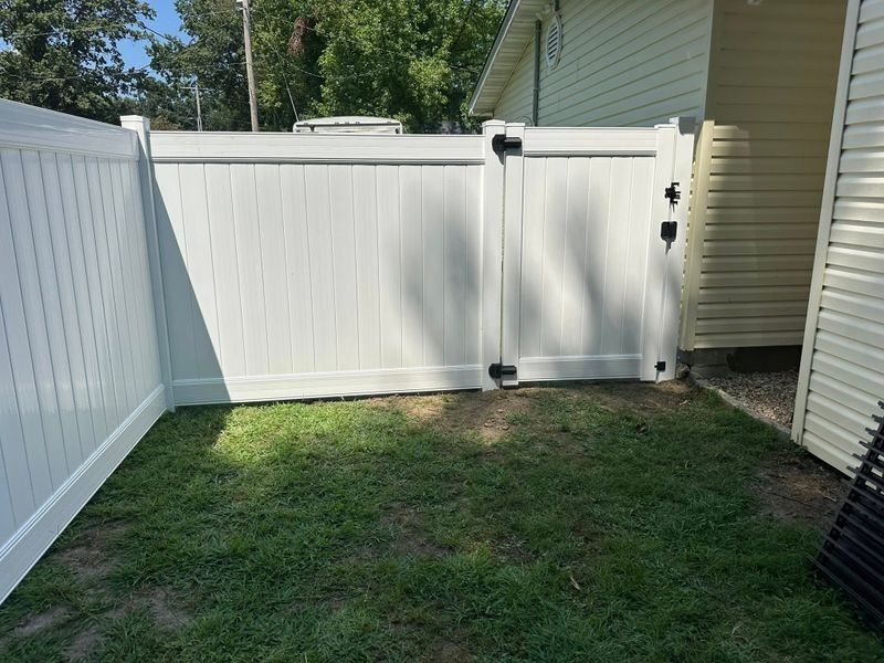 Small fenced backyard with grass, white vinyl fence and gate beside a house in daylight