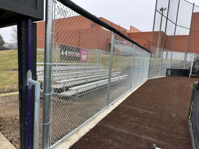 Empty bleachers beside a fenced sports field with red-roofed buildings in the background