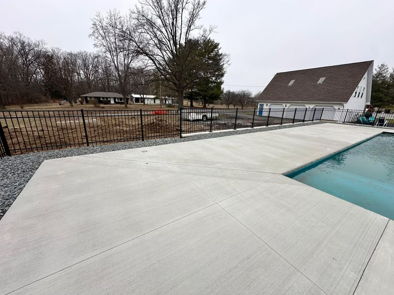 Concrete pool deck beside turquoise swimming pool with a fenced yard and a house in the background