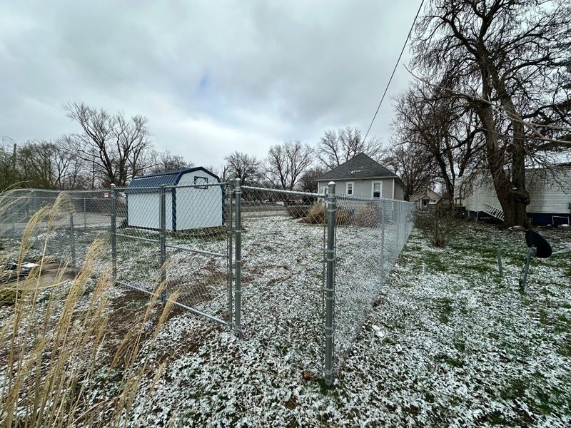Snow-dusted fenced yard with a small shed and house under a cloudy sky