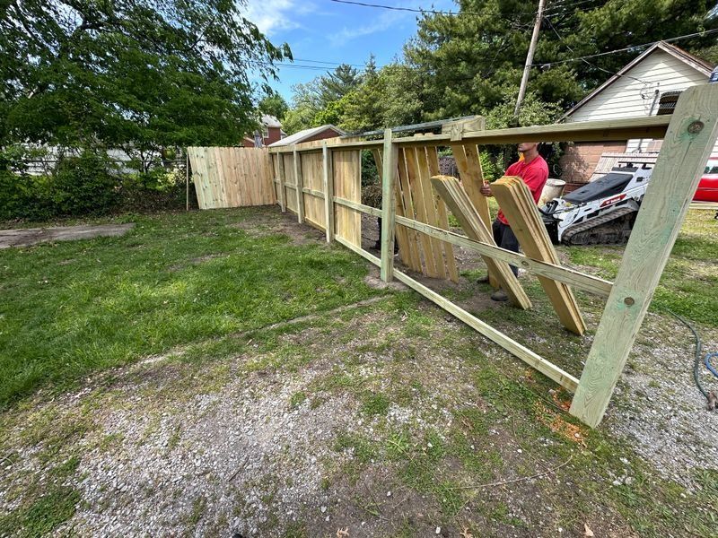 Workers building a wooden fence along a grassy driveway beside a house and parked cars