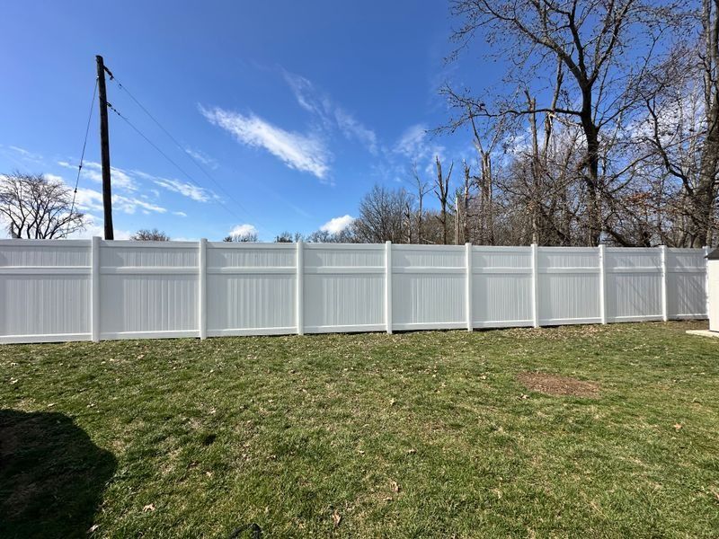 White vinyl fence across a grassy yard under a blue sky, with trees and utility poles in the background