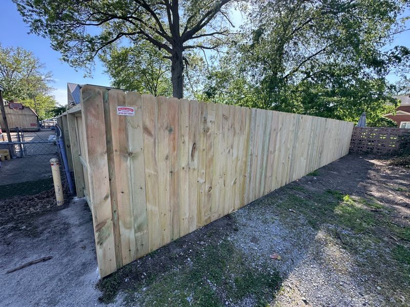 Long wooden privacy fence along a gravel yard with trees and a brick wall behind it