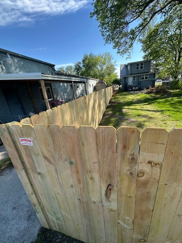 Wooden fence along a driveway beside houses and a grassy yard under a blue sky