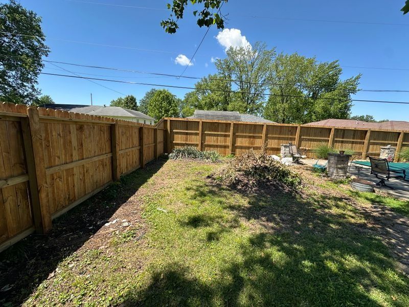 Backyard with wooden privacy fence, patchy grass, and a pile of yard debris under a bright blue sky
