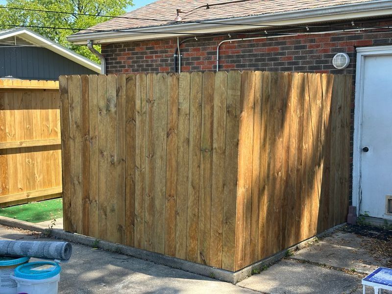 Wooden privacy fence enclosure beside a brick house, with a side gate and concrete patio.