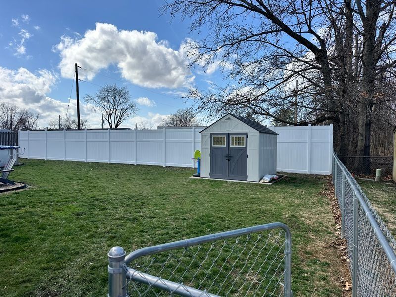 Backyard with white privacy fence, small gray shed, chain-link gate, and grassy yard under a cloudy sky