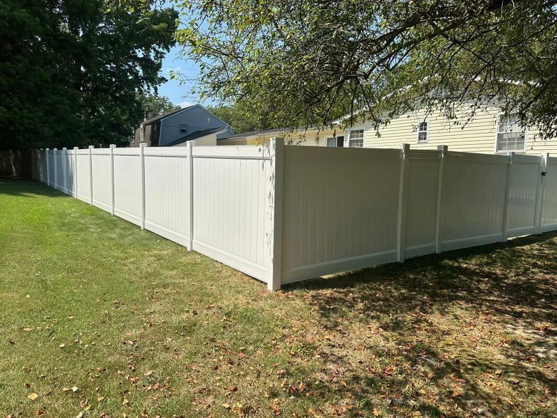 White vinyl fence running along a grassy yard beside a house, under trees in daylight