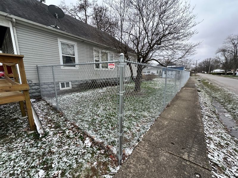 Snow-dusted chain-link fence beside a gray house along a sidewalk on an overcast street.