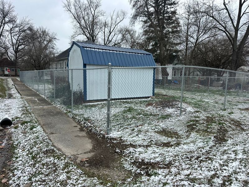 Snow-dusted fenced yard with a small blue shed and a concrete path under bare trees