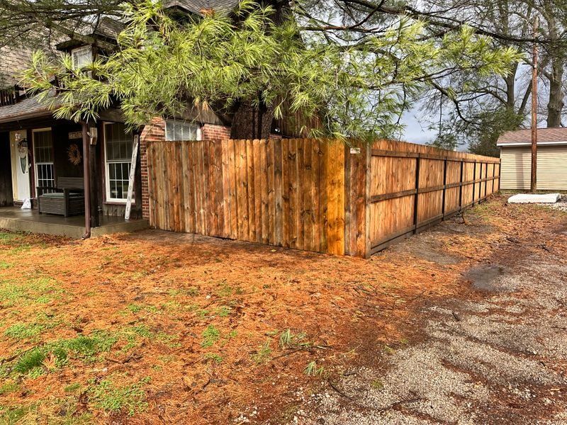 Wooden privacy fence beside a house, with leafy trees and a gravel driveway in a yard.