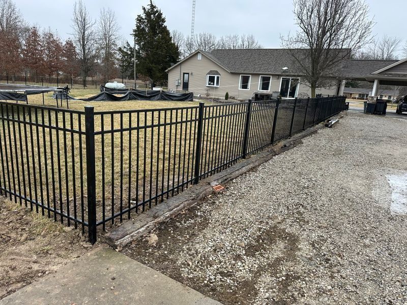 Black metal fence around a yard beside a gravel driveway and beige house on an overcast day