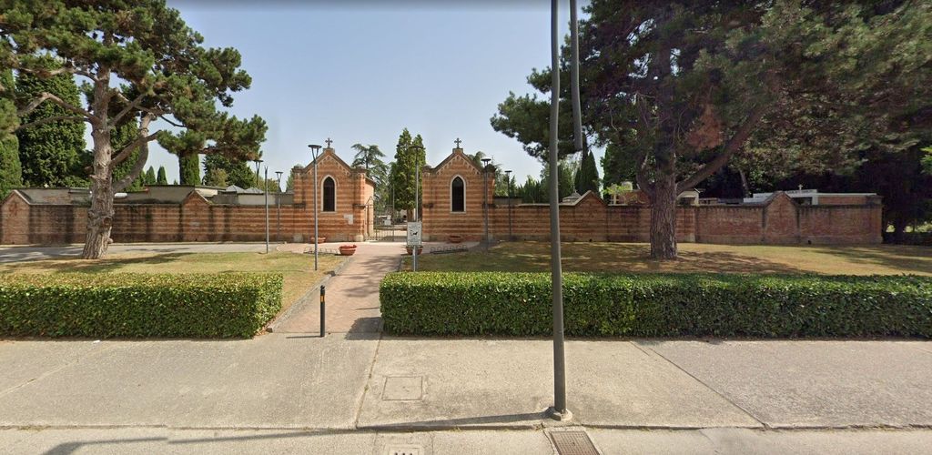 Cappella in mattoni con ingresso ad arco, piccola finestra rotonda e alti cipressi lungo un muro di mattoni.