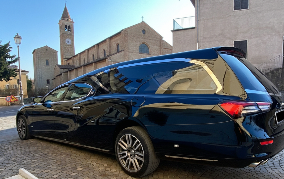 Un carro funebre nero parcheggiato su una strada acciottolata, con una chiesa sullo sfondo in una giornata di sole.