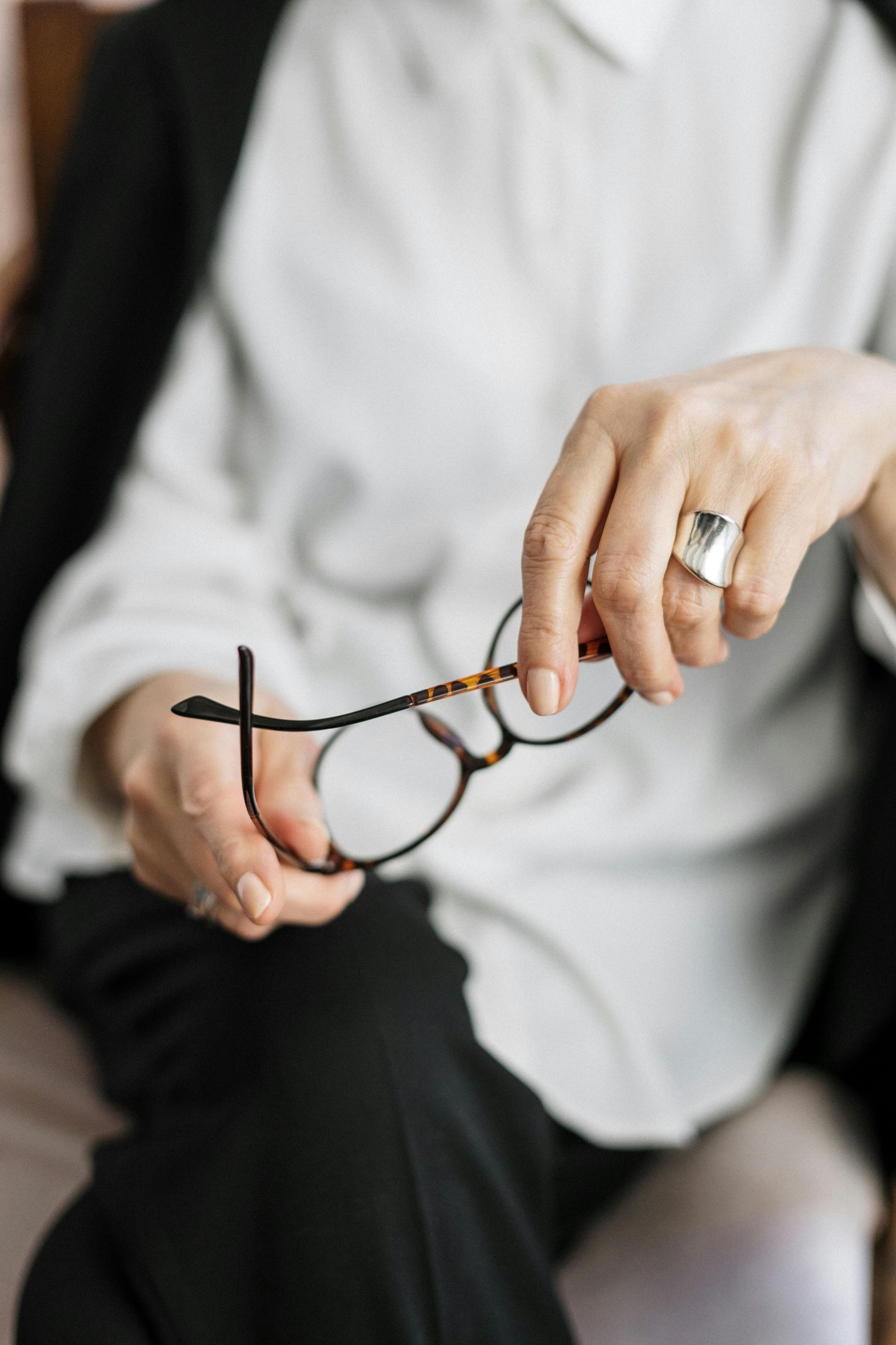 Person holding eyeglasses, wearing a white shirt and black blazer. A ring is visible on their finger.