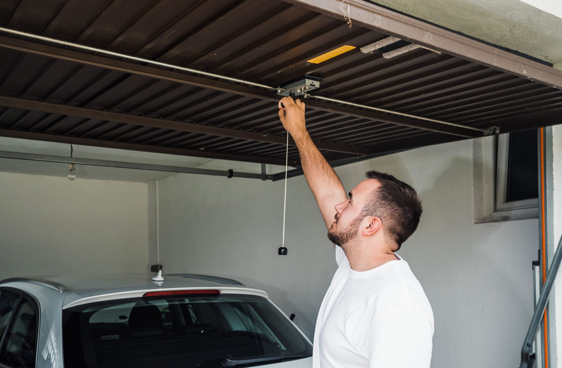 Man in garage reaching up to garage door opener. Car visible.