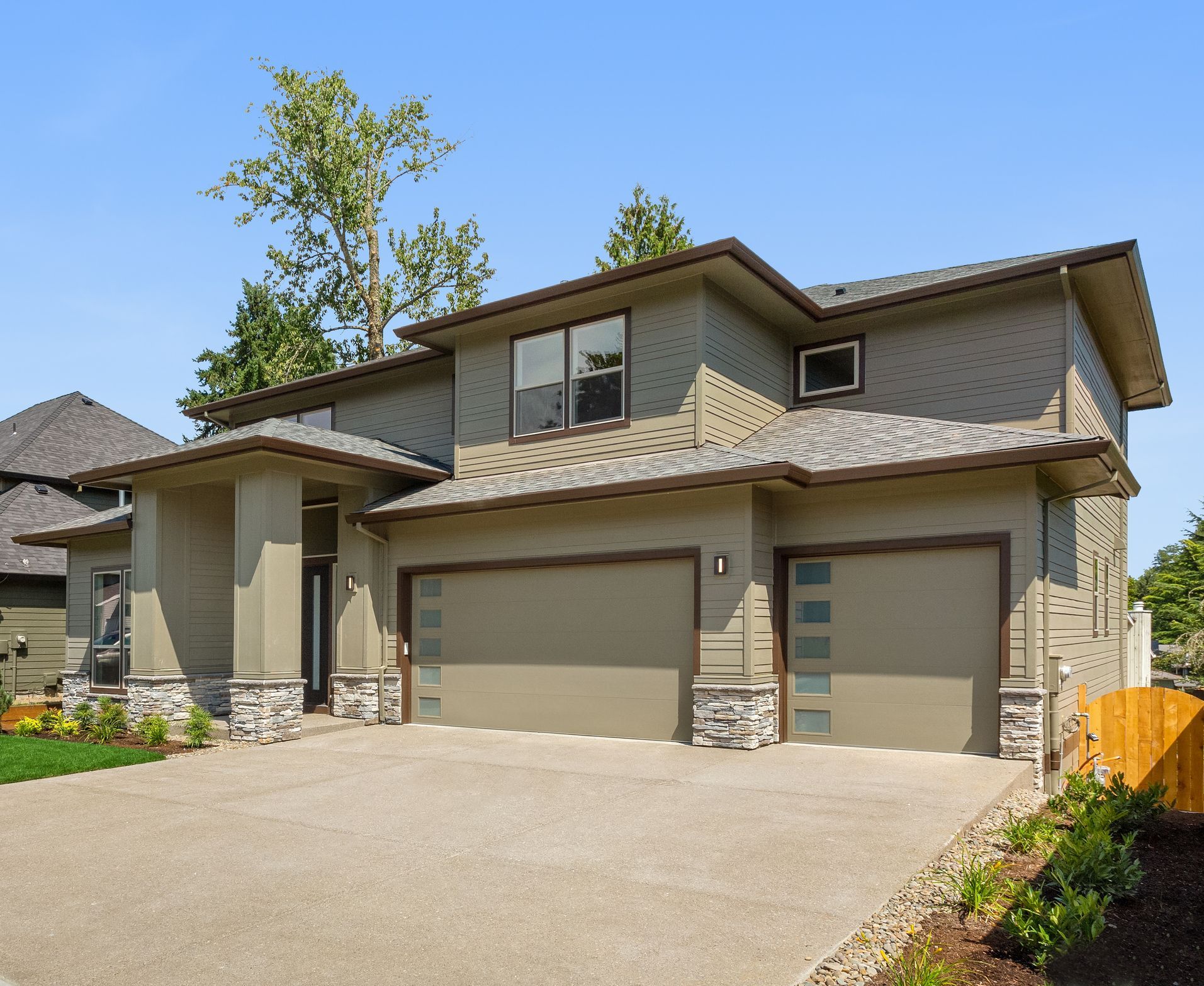 Modern two-story house with two-car garage. Tan siding, stone accents, and concrete driveway under blue sky.