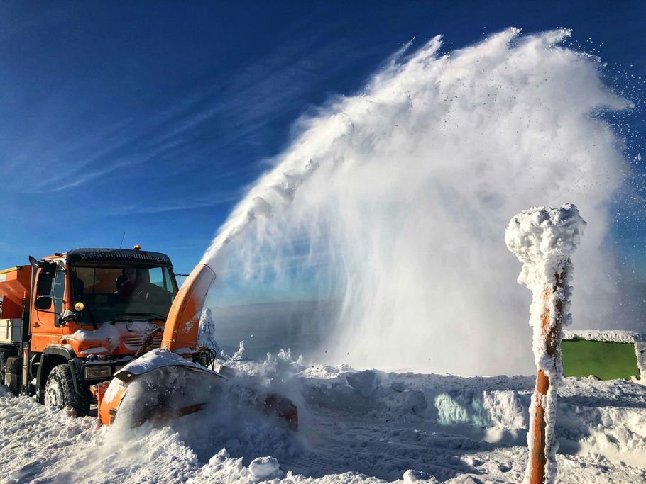 Orange snowplow clearing heavy snow with a large spray on a road under a blue sky