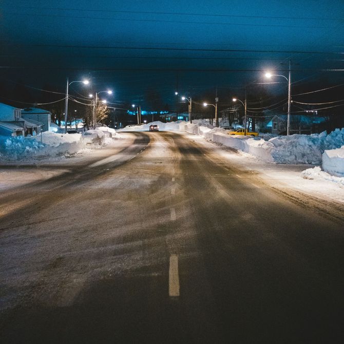 Snow-covered road at night under streetlights, with tire tracks and houses on both sides.