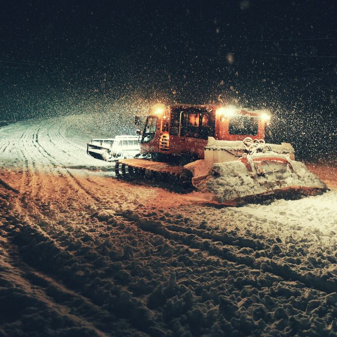 Snowplow clearing a snow-covered road at night, with orange lights glowing in falling snow.