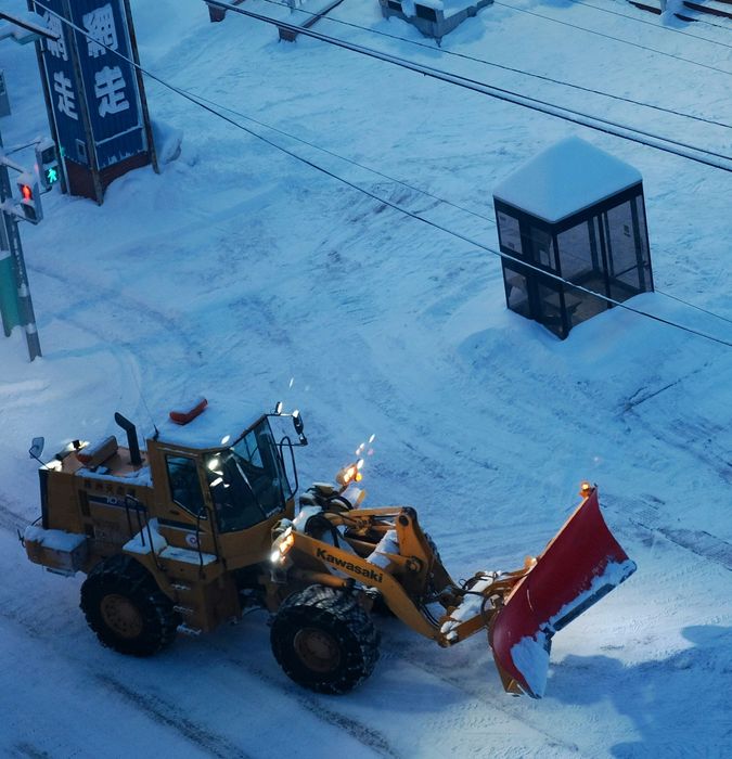Yellow snowplow clearing snow on a snowy street near a bus stop and traffic lights