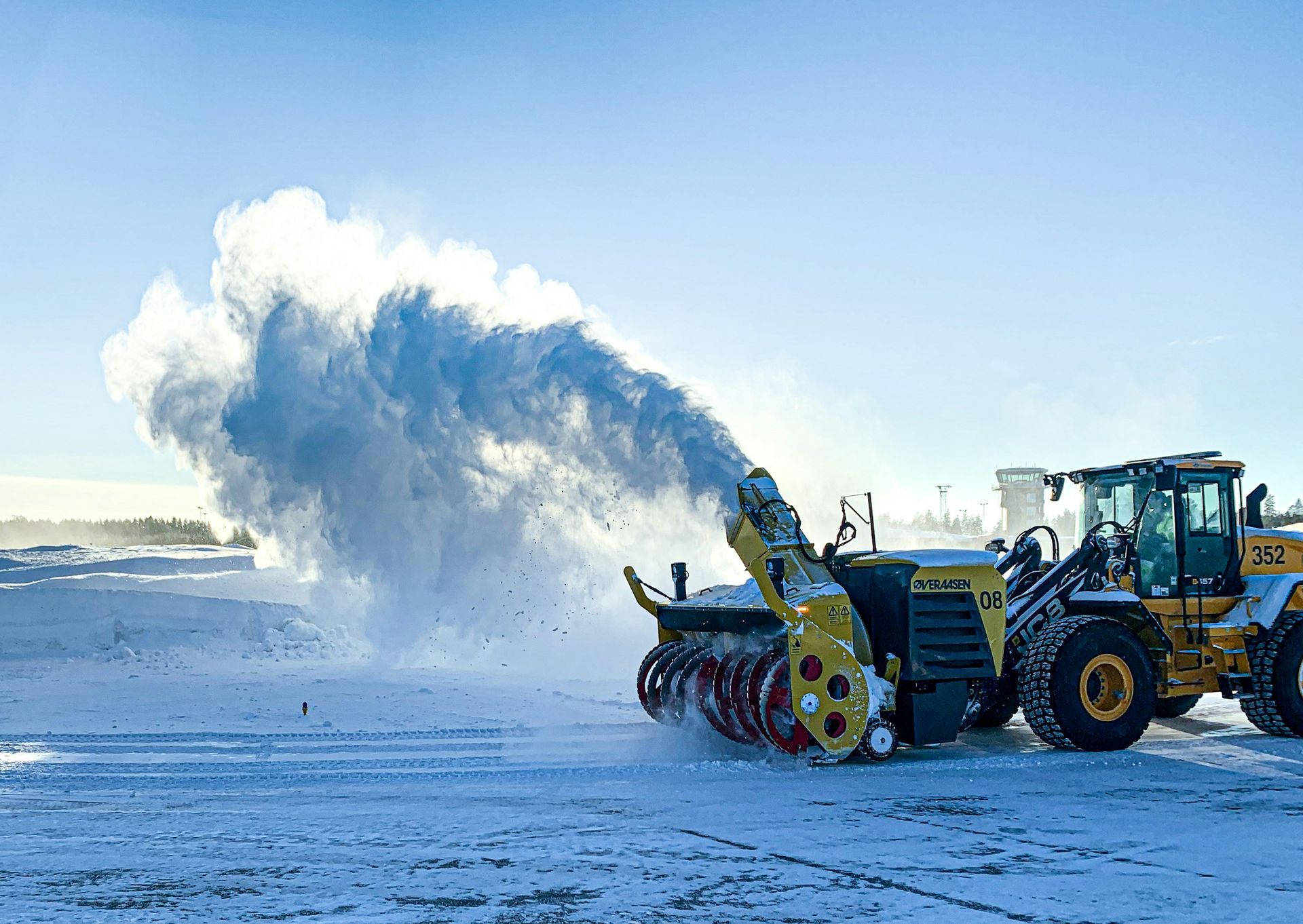 Snowplow clearing heavy snow, blasting a plume across a snowy road under a blue sky.