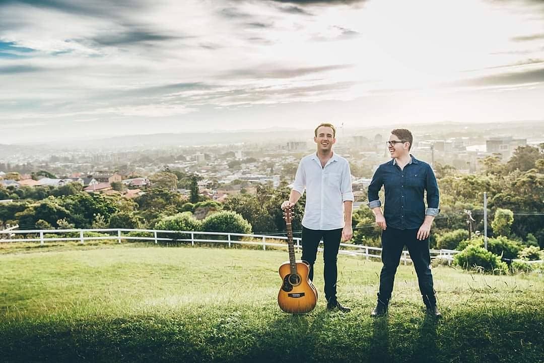 Two men are standing in a field holding guitars.