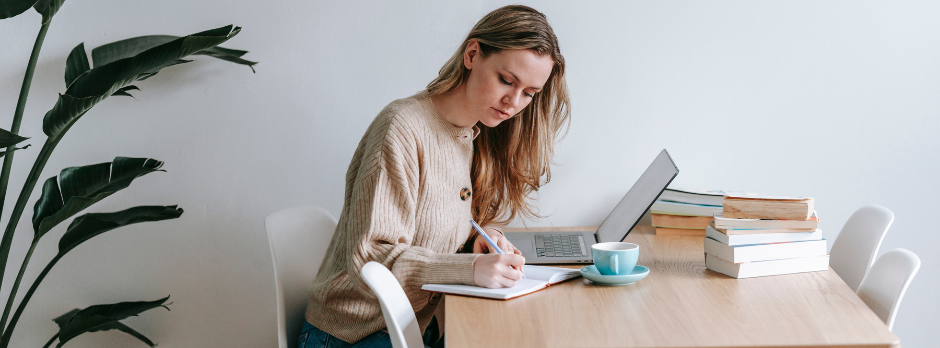 A woman is sitting at a table with a laptop and writing in a notebook.