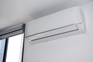 Wall-mounted white air conditioner beneath a window with a black blind