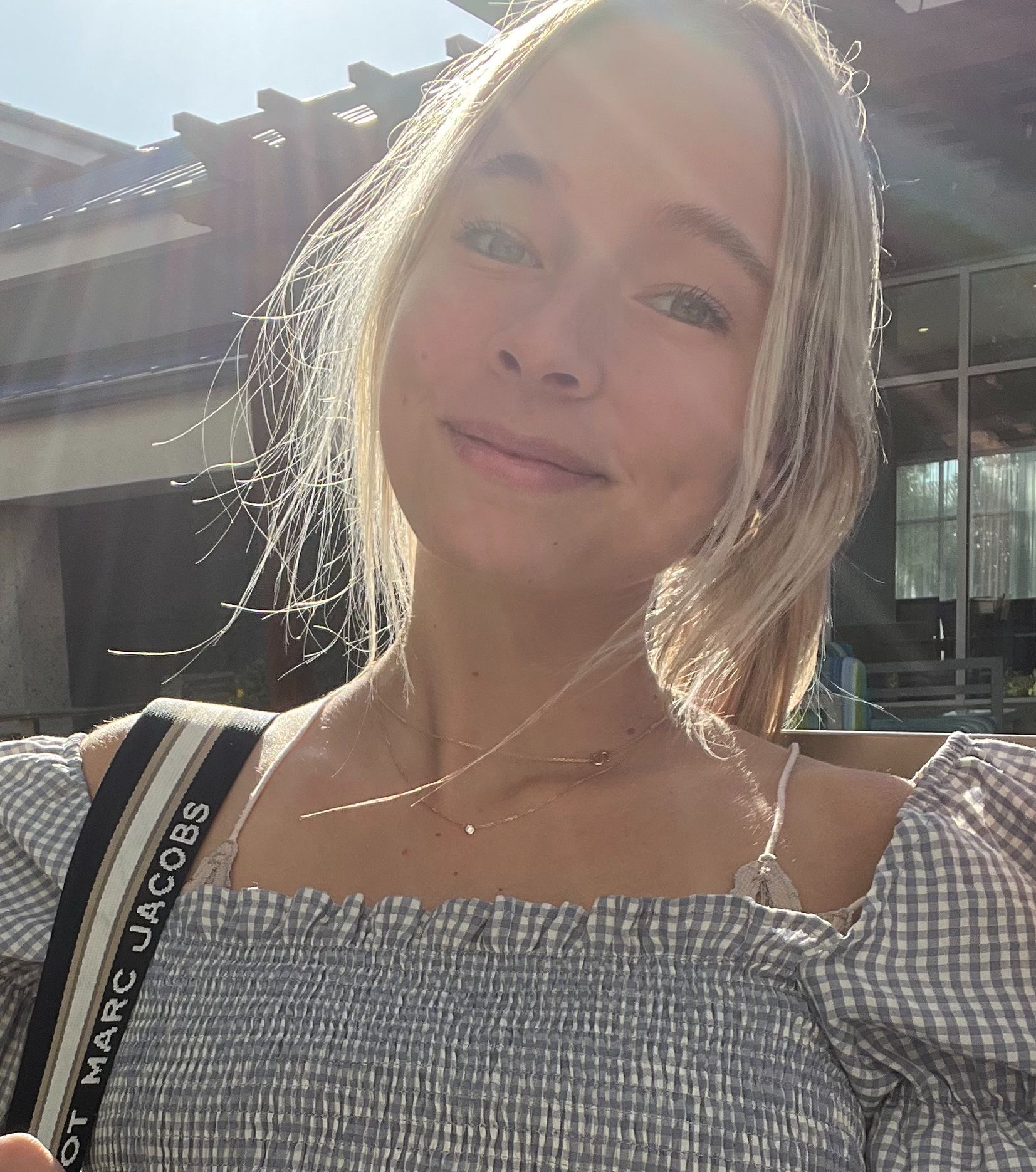 Smiling woman in a gray top outdoors in sunlight, with a Marc Jacobs strap visible over her shoulder.