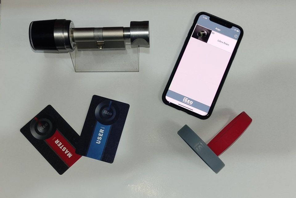 Un cilindro di serratura intelligente, due telecomandi RFID e uno smartphone che visualizza un'app per il controllo della serratura su una superficie bianca.