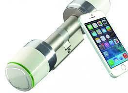 Un cilindro di serratura intelligente in metallo con una manopola elettronica bianca, abbinato a uno smartphone che visualizza l'interfaccia di un'app.
