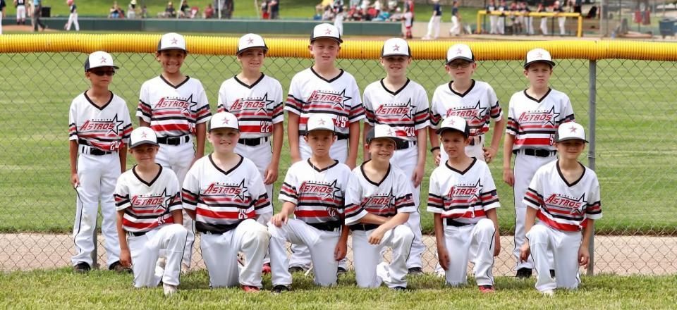 Youth baseball team posing in uniform on a field in front of a fence and trees
