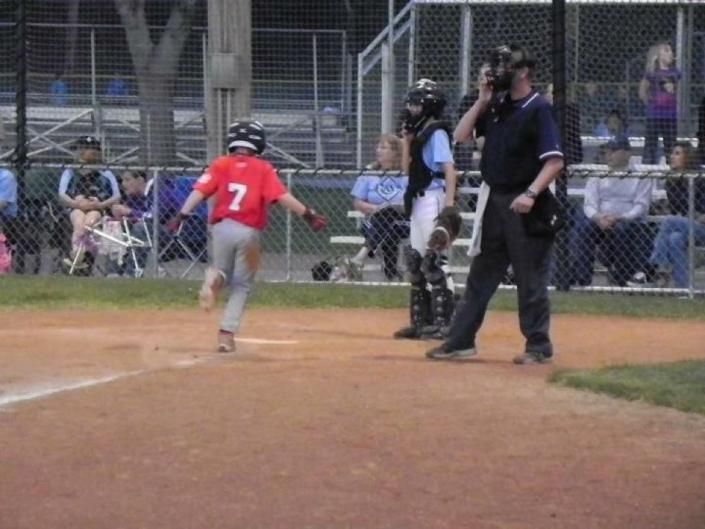 Youth baseball player in red jersey runs toward home plate as umpire and catcher stand nearby.
