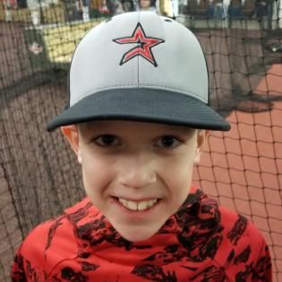 Child in a red shirt and gray cap smiling in front of a baseball net
