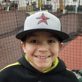 Smiling child in a black hoodie and white cap with red star, standing in front of a net at an indoor sports facility