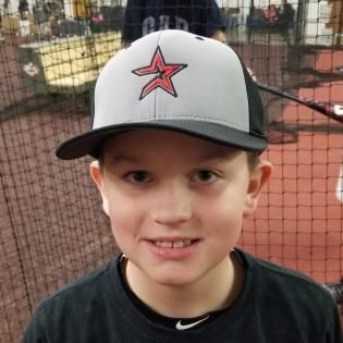 Smiling child in a gray cap with a red star logo, standing in front of a netted sports backdrop