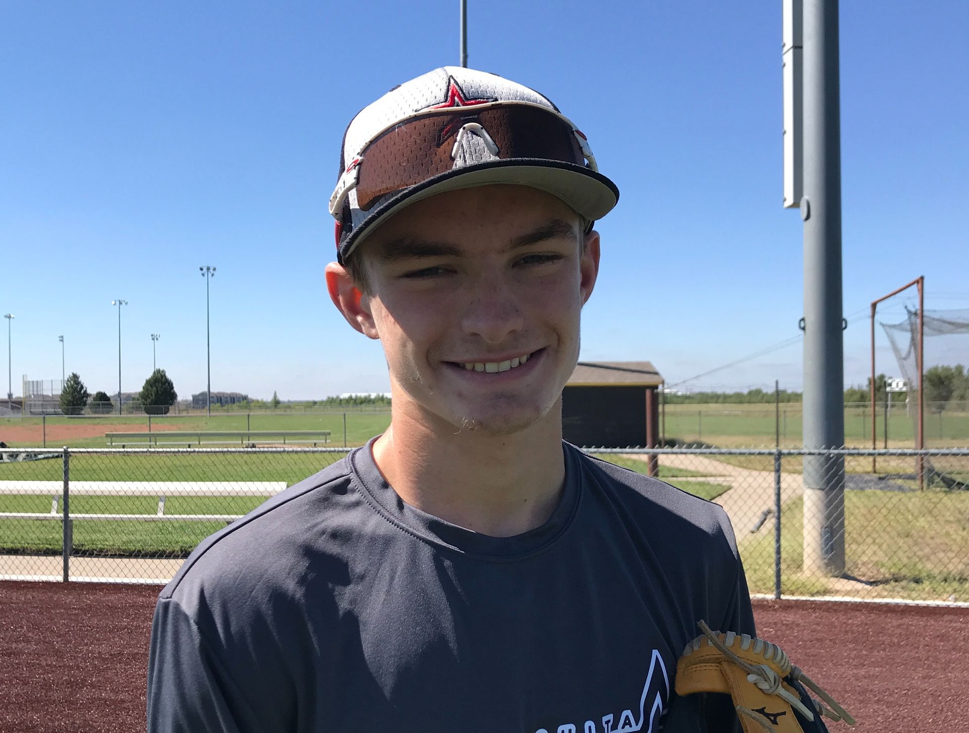 Smiling baseball player in a cap and dark shirt on a field, holding a glove under a clear blue sky