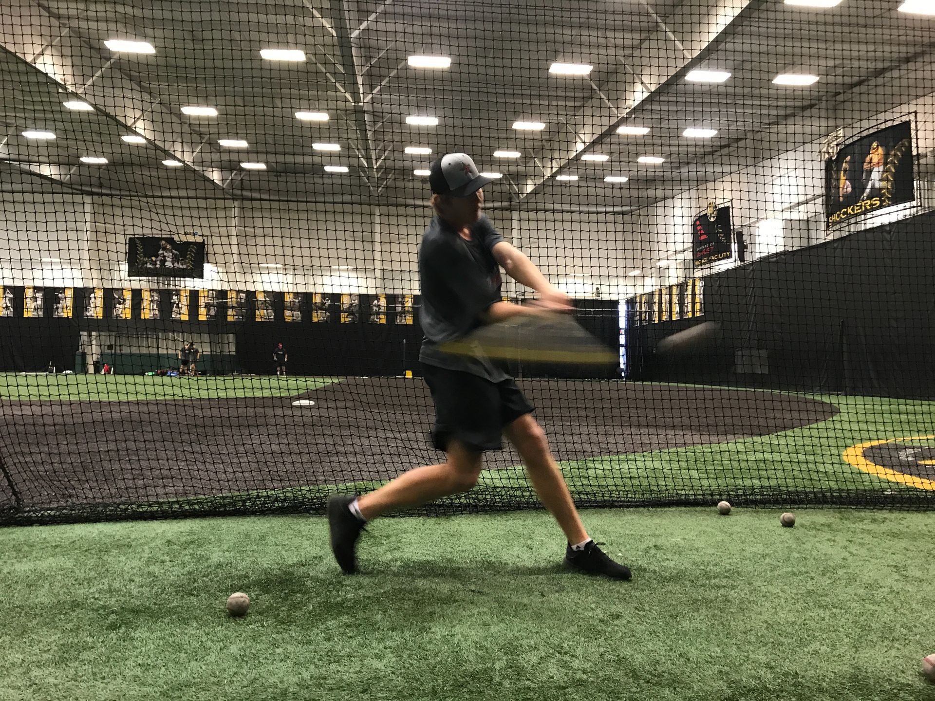Indoor batting cage with a player swinging a bat on green turf behind a net.
