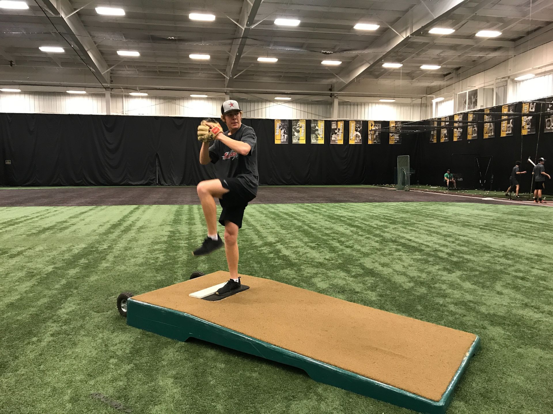 Baseball pitcher in black training on an indoor mound, mid-leg lift, inside a turf facility.