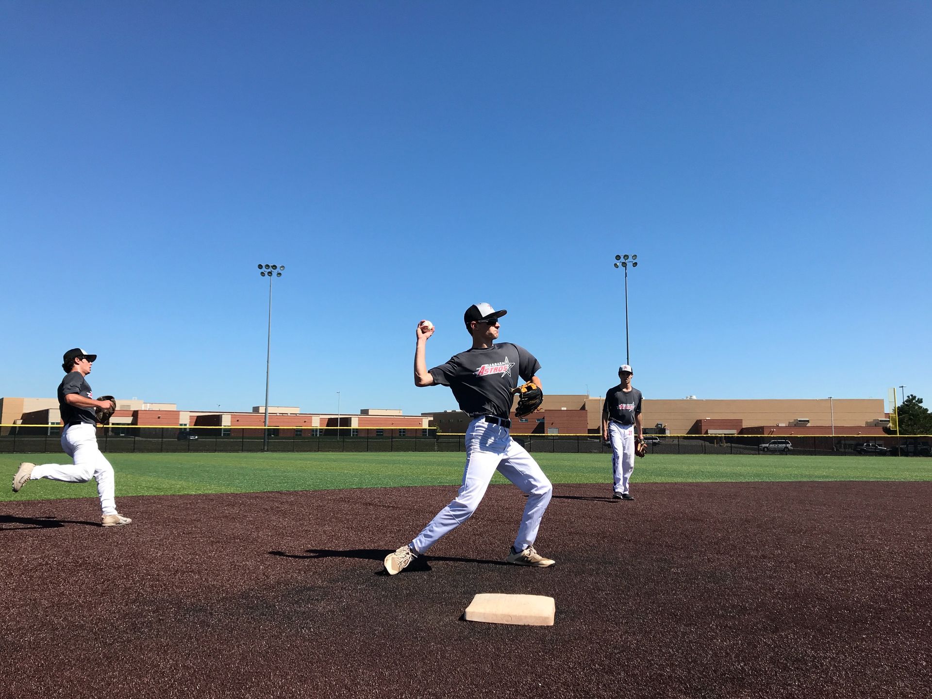 Baseball pitcher on the mound during practice, with two teammates nearby on a field under a clear blue sky.
