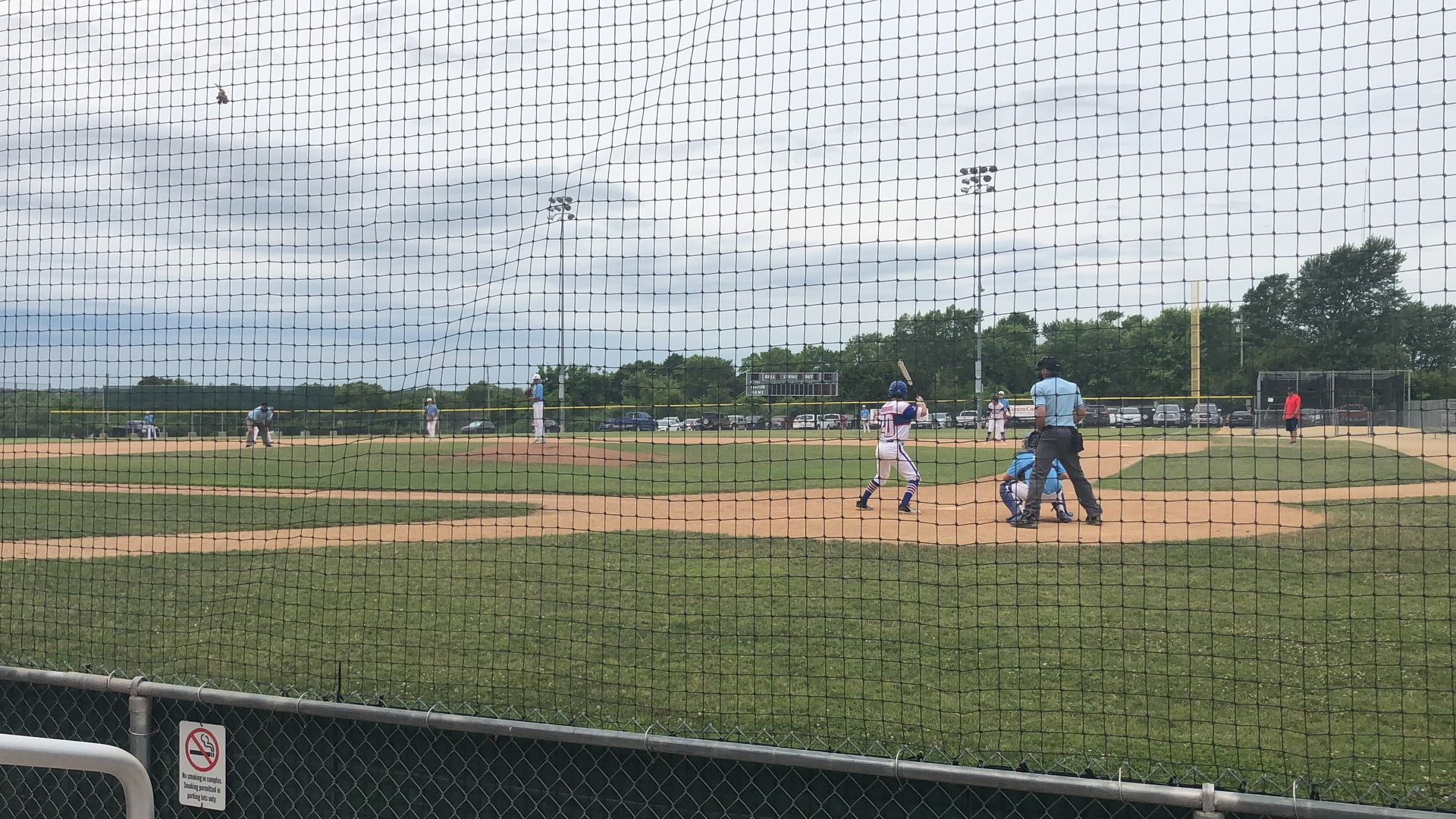 Baseball game in progress at a field, viewed through protective netting under a cloudy sky.