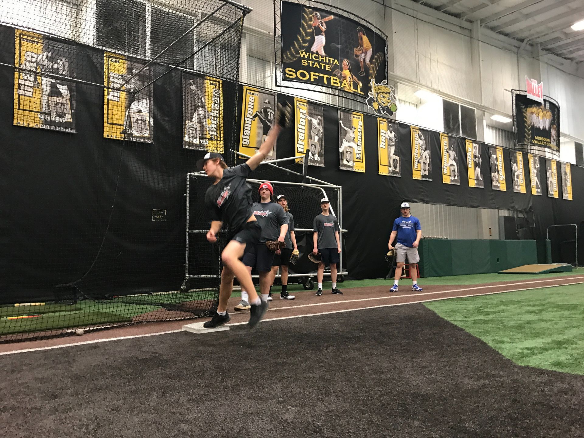 Athletes running drills on an indoor turf track near a netted wall and hanging banners