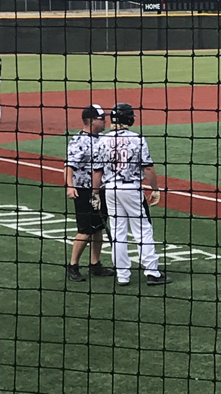 Two baseball players in white uniforms talking on the field behind a net.