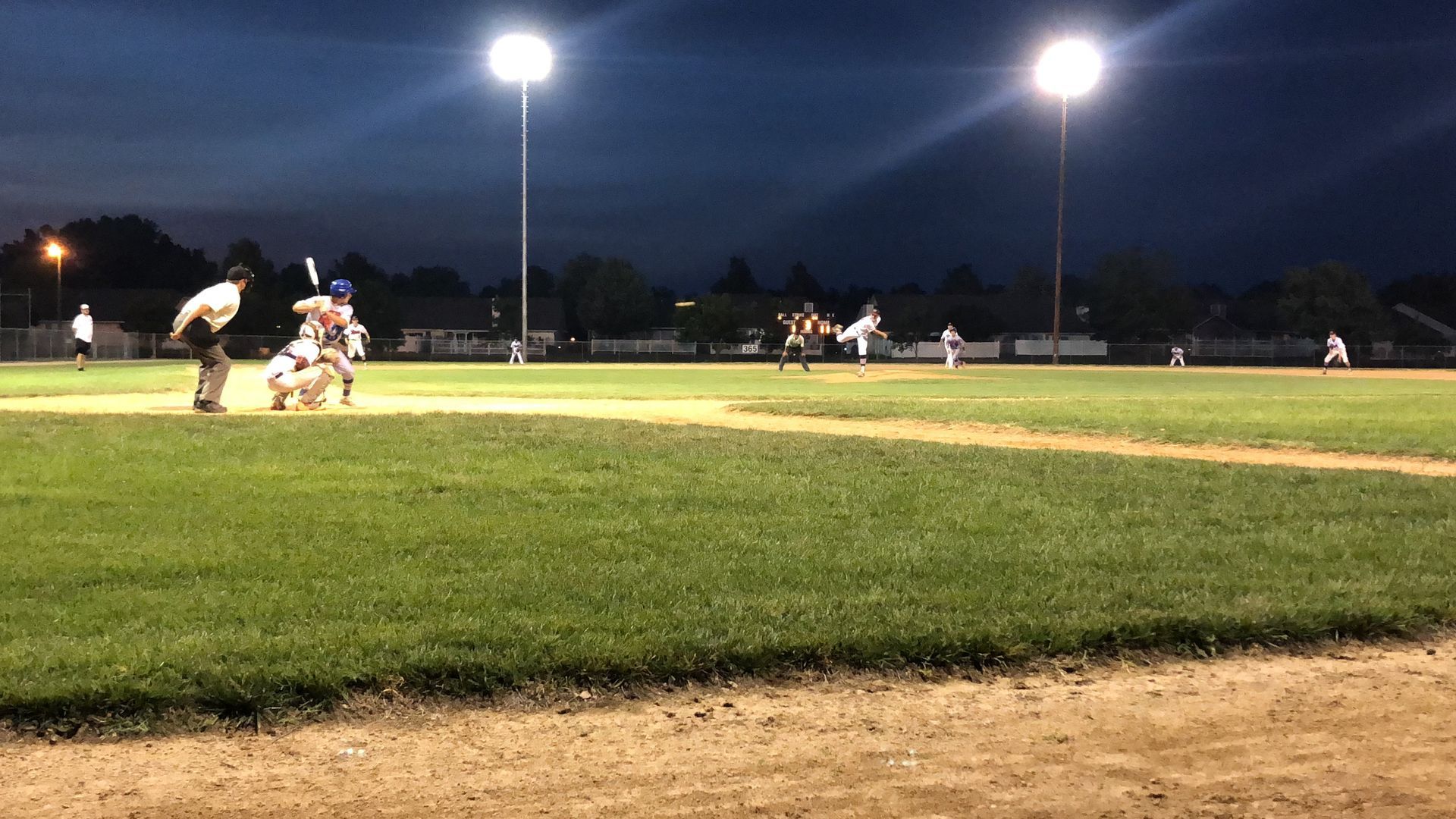 Nighttime baseball game under bright field lights, with players on the field and a batter at home plate.