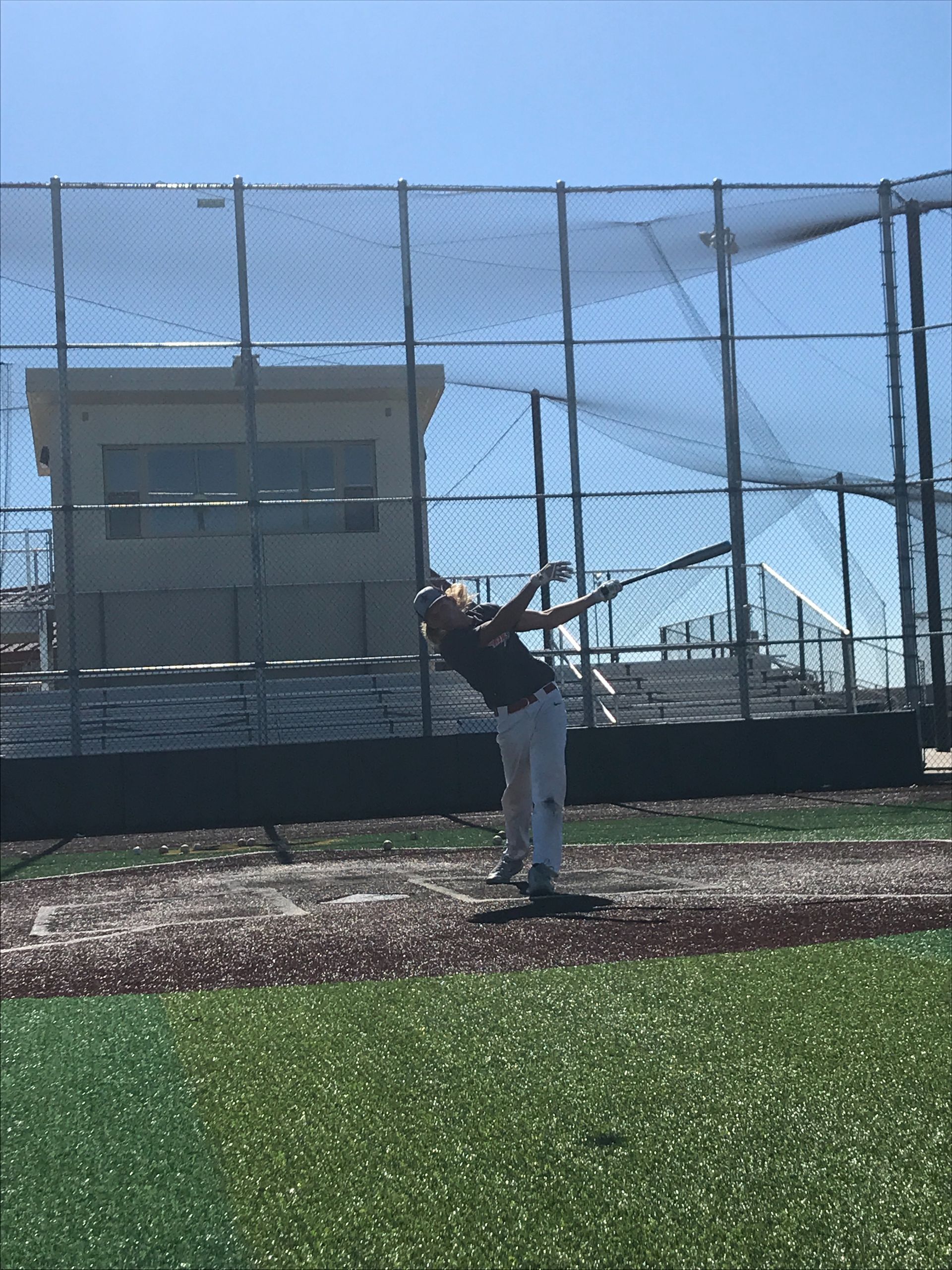 Baseball player mid-swing on a turf field near a chain-link fence and building
