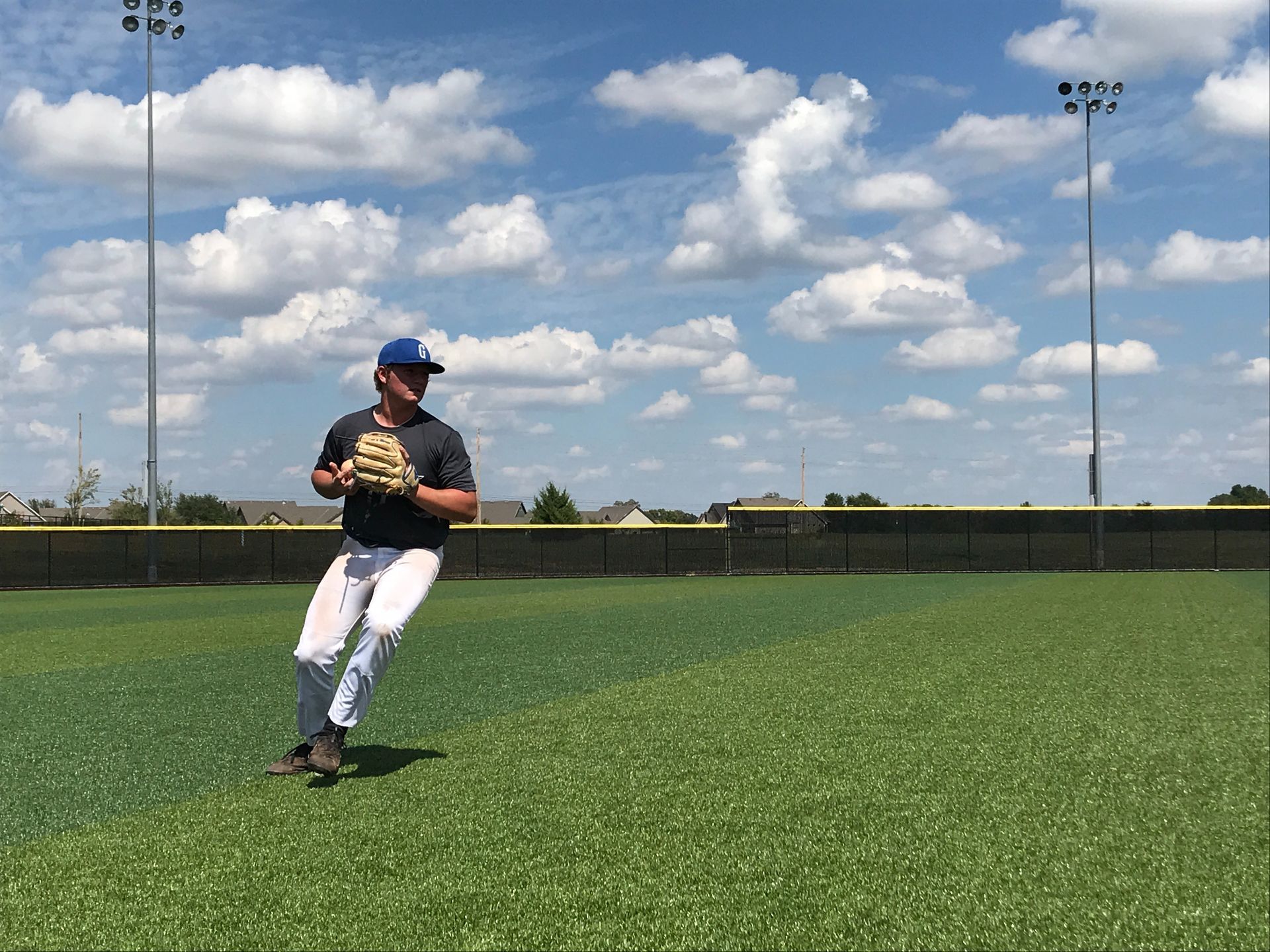 Baseball player in black jersey and white pants fielding a ball on a grassy field under a blue sky.
